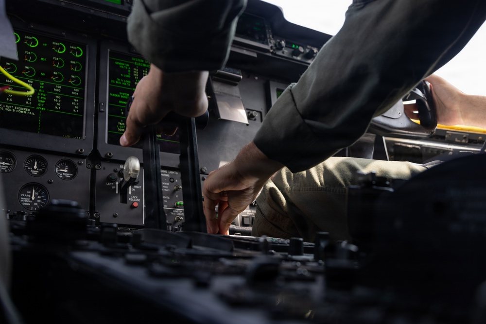 The image shows the interior cockpit of a military aircraft, with two individuals in flight suits operating controls and instrumentation panels. Multiple green-lit digital displays and analog gauges are visible, suggesting an active pre-flight or in-flight checklist procedure. The low-angle, close-up perspective emphasizes the complexity of the cockpit environment.