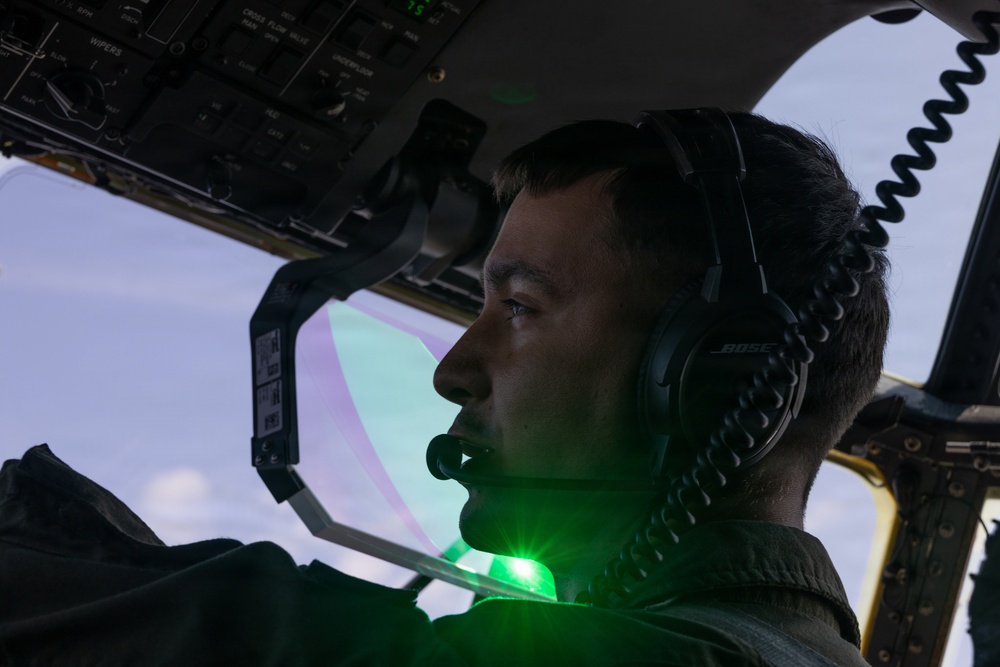 A pilot or aircrew member is seated in a military or large aircraft cockpit, wearing a Bose aviation headset with a boom microphone. A bright green light source is visible near the lower portion of the frame, casting a green glow on the subject's chin and neck area. The cockpit overhead panel with labeled switches and controls is visible in the background.