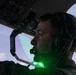 A pilot or aircrew member is seated in a military or large aircraft cockpit, wearing a Bose aviation headset with a boom microphone. A bright green light source is visible near the lower portion of the frame, casting a green glow on the subject's chin and neck area. The cockpit overhead panel with labeled switches and controls is visible in the background.