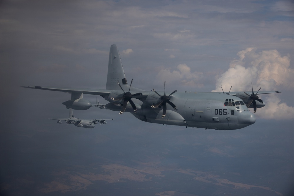 The image shows multiple military turboprop transport aircraft flying in formation at altitude. The lead aircraft, marked with the number '065', is a C-130 Hercules variant in a gray military paint scheme. At least two additional C-130-type aircraft are visible in the background flying in loose formation.