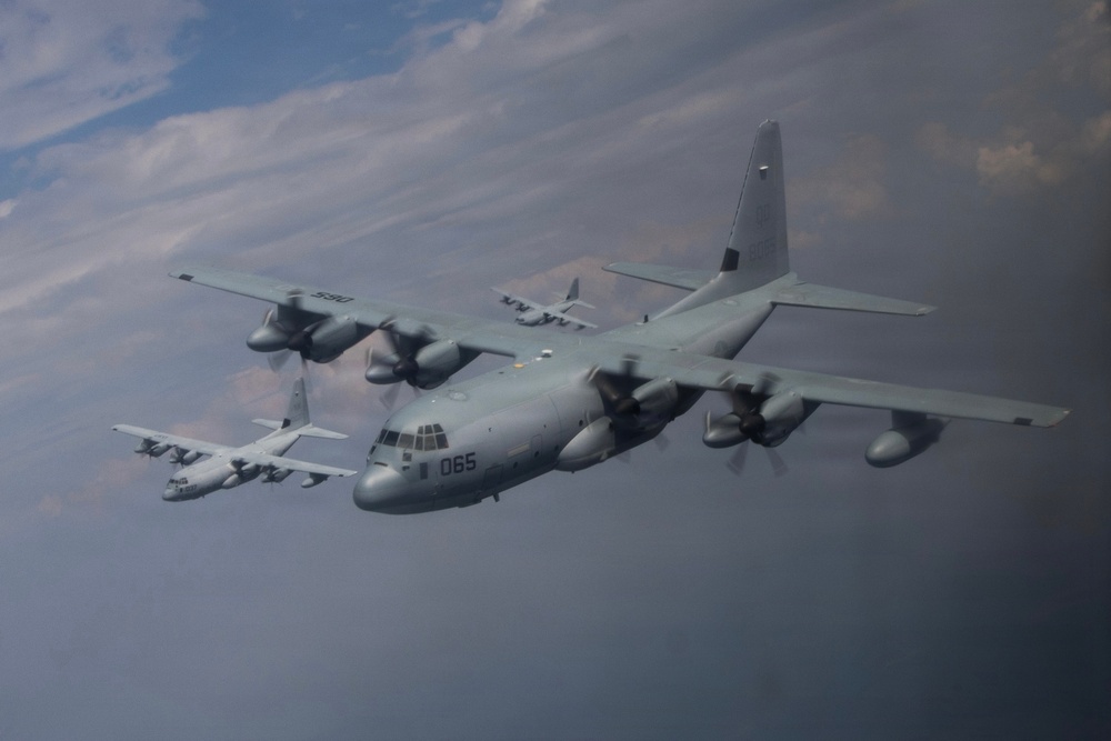 The image shows a formation flight of multiple C-130 Hercules turboprop military transport aircraft flying in close proximity against a partly cloudy sky. The foreground aircraft bears the number '065' on its nose, while a second aircraft marked '590' is visible behind and to the left, with at least one additional aircraft visible in the background. The aircraft appear to be in a gray military livery consistent with U.S. Marine Corps or U.S. Navy markings.