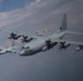 The image shows a formation flight of multiple C-130 Hercules turboprop military transport aircraft flying in close proximity against a partly cloudy sky. The foreground aircraft bears the number '065' on its nose, while a second aircraft marked '590' is visible behind and to the left, with at least one additional aircraft visible in the background. The aircraft appear to be in a gray military livery consistent with U.S. Marine Corps or U.S. Navy markings.