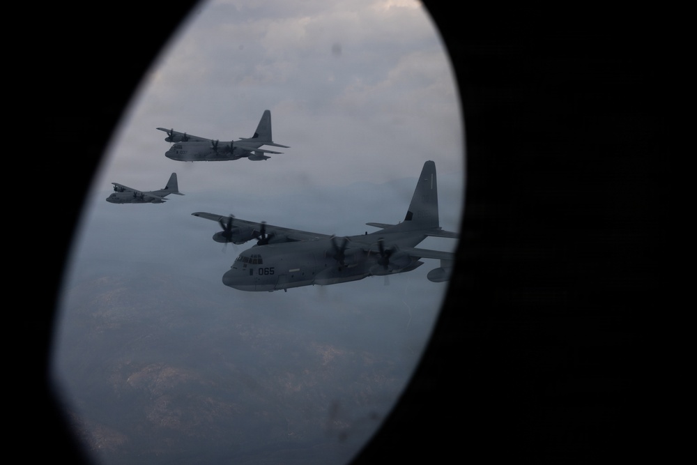 The image is taken through an oval aircraft porthole window, showing a formation of four military turboprop transport aircraft flying in formation. The closest aircraft bears the visible tail number '065' and is identifiable as a C-130 Hercules variant. The aircraft are flying at altitude over a hazy, partially cloud-covered landscape.