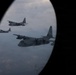 The image is taken through an oval aircraft porthole window, showing a formation of four military turboprop transport aircraft flying in formation. The closest aircraft bears the visible tail number '065' and is identifiable as a C-130 Hercules variant. The aircraft are flying at altitude over a hazy, partially cloud-covered landscape.