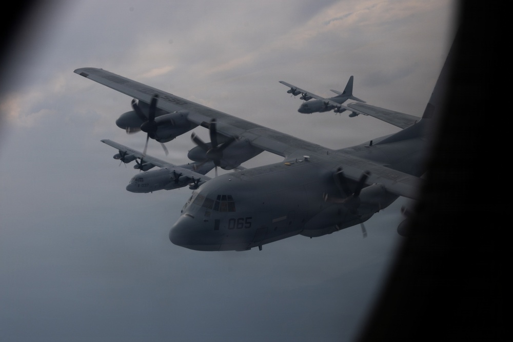 The image shows a formation flight of multiple C-130 Hercules military transport aircraft photographed through a window of another aircraft. At least four C-130s are visible in close formation against an overcast sky. The nearest aircraft bears the tail number '065' and appears to be a US Marine Corps variant.