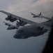 The image shows a formation flight of multiple C-130 Hercules military transport aircraft photographed through a window of another aircraft. At least four C-130s are visible in close formation against an overcast sky. The nearest aircraft bears the tail number '065' and appears to be a US Marine Corps variant.