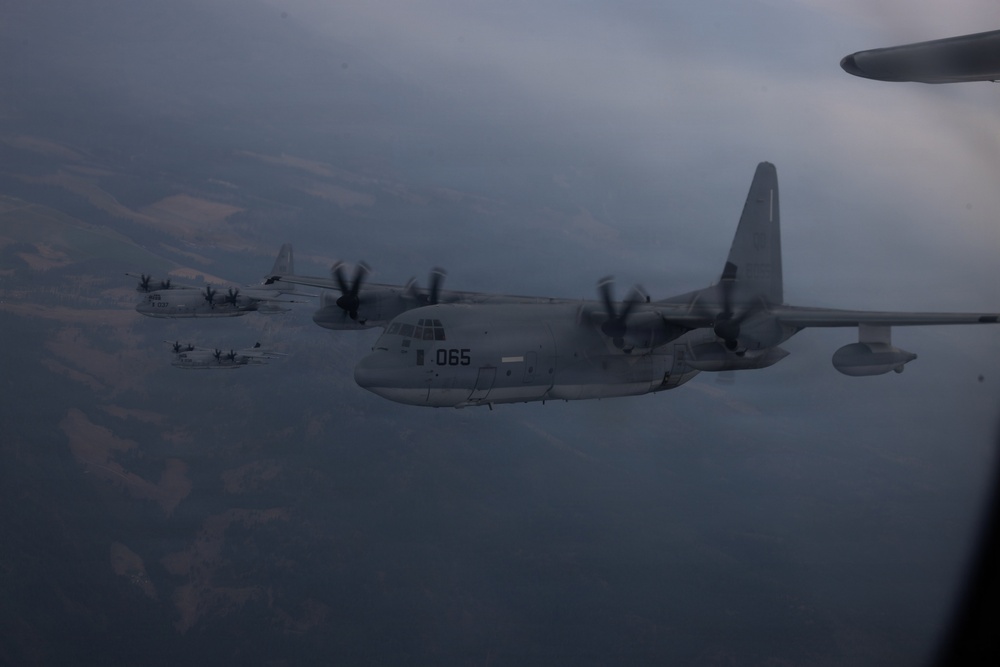 A formation of military turboprop transport aircraft, identifiable as C-130 Hercules variants, flying in formation over mountainous terrain under overcast skies. The lead aircraft bears the visible tail number '065' and appears to be a US Marine Corps KC-130 tanker variant, equipped with underwing refueling pods. The image is taken from another aircraft, with a partial wing or fuselage visible in the upper right corner.