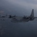 A formation of military turboprop transport aircraft, identifiable as C-130 Hercules variants, flying in formation over mountainous terrain under overcast skies. The lead aircraft bears the visible tail number '065' and appears to be a US Marine Corps KC-130 tanker variant, equipped with underwing refueling pods. The image is taken from another aircraft, with a partial wing or fuselage visible in the upper right corner.