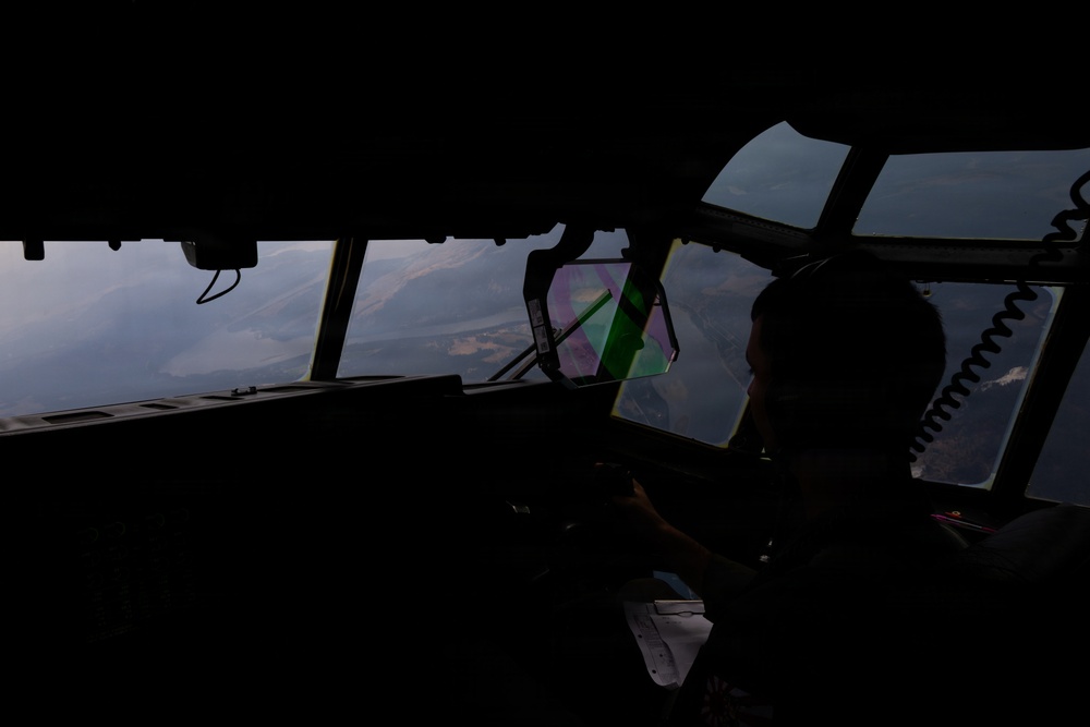 A silhouetted pilot is seated in the cockpit of a large military or transport aircraft, flying over mountainous terrain. The interior is heavily shadowed with the exterior landscape visible through multiple cockpit windows. Hazy or smoky atmospheric conditions are visible outside, suggesting possible wildfire smoke or low visibility weather.