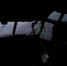 A silhouetted pilot is seated in the cockpit of a large military or transport aircraft, flying over mountainous terrain. The interior is heavily shadowed with the exterior landscape visible through multiple cockpit windows. Hazy or smoky atmospheric conditions are visible outside, suggesting possible wildfire smoke or low visibility weather.