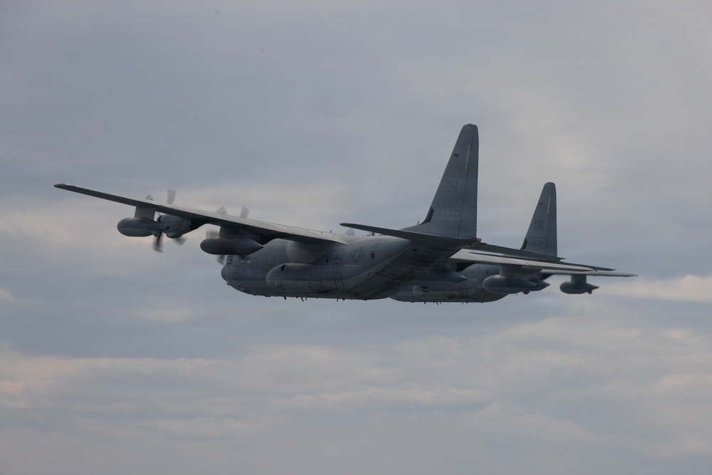 A military turboprop transport aircraft, consistent with a Lockheed C-130 Hercules variant, is photographed in flight against an overcast sky. The aircraft is painted in a flat gray military finish and appears to be in level flight or a slight bank. Four turboprop engines with spinning propellers are visible, and the aircraft shows various external pods and antennas suggesting a specialized variant.