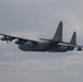A military turboprop transport aircraft, consistent with a Lockheed C-130 Hercules variant, is photographed in flight against an overcast sky. The aircraft is painted in a flat gray military finish and appears to be in level flight or a slight bank. Four turboprop engines with spinning propellers are visible, and the aircraft shows various external pods and antennas suggesting a specialized variant.