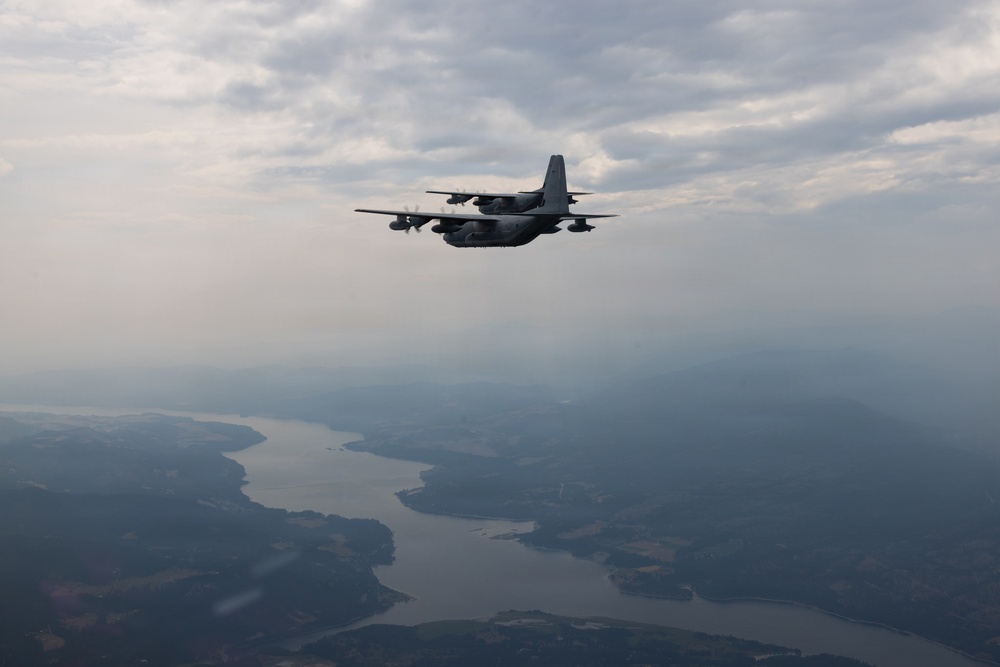 A military turboprop aircraft, consistent with a C-130 Hercules variant, is photographed in flight from an air-to-air perspective. The aircraft is flying over a landscape featuring a winding river or reservoir system surrounded by forested terrain. The sky is overcast with scattered clouds and hazy atmospheric conditions.