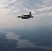A military turboprop aircraft, consistent with a C-130 Hercules variant, is photographed in flight from an air-to-air perspective. The aircraft is flying over a landscape featuring a winding river or reservoir system surrounded by forested terrain. The sky is overcast with scattered clouds and hazy atmospheric conditions.