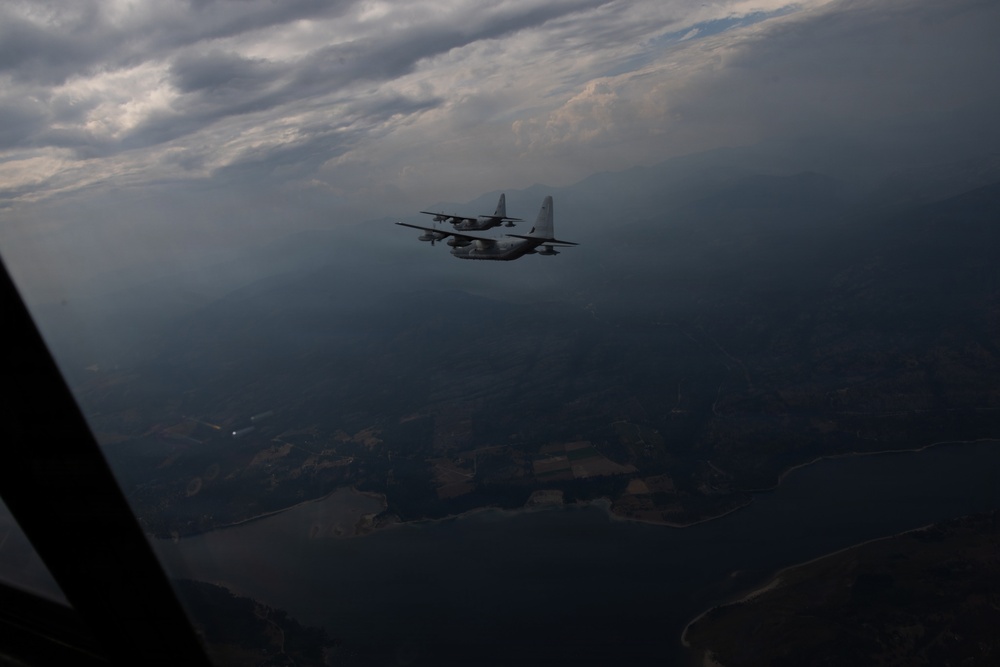 Two military turboprop transport aircraft, consistent with C-130 Hercules variants, are flying in close formation at altitude. The photo is taken from a third aircraft, visible by the window frame in the lower left corner. The scene is dramatic with overcast, moody skies and a mountainous, partially forested landscape visible below.