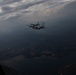 Two military turboprop transport aircraft, consistent with C-130 Hercules variants, are flying in close formation at altitude. The photo is taken from a third aircraft, visible by the window frame in the lower left corner. The scene is dramatic with overcast, moody skies and a mountainous, partially forested landscape visible below.