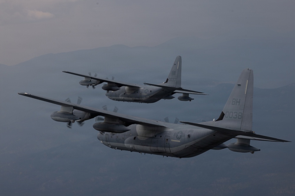 Two military turboprop transport aircraft are flying in formation over a mountainous, hazy landscape. Both aircraft are painted in overall gray camouflage and bear U.S. military markings consistent with the C-130 Hercules platform. The foreground aircraft displays tail code 'BH' and serial number '0038', while the background aircraft shows serial number '0037'.