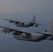 Two military turboprop transport aircraft are flying in formation over a mountainous, hazy landscape. Both aircraft are painted in overall gray camouflage and bear U.S. military markings consistent with the C-130 Hercules platform. The foreground aircraft displays tail code 'BH' and serial number '0038', while the background aircraft shows serial number '0037'.
