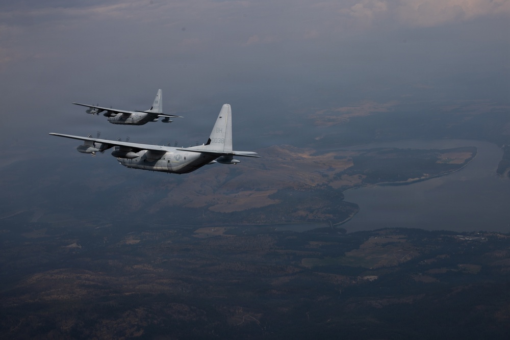 Two military transport aircraft, identifiable as C-130 Hercules variants, are flying in formation over a landscape featuring rivers, forests, and rolling terrain. The foreground aircraft displays tail markings including 'BH' and what appears to be '038' or similar serial numbers. The image is taken from another aircraft at altitude during what appears to be dusk or overcast conditions.