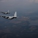 Two military transport aircraft, identifiable as C-130 Hercules variants, are flying in formation over a landscape featuring rivers, forests, and rolling terrain. The foreground aircraft displays tail markings including 'BH' and what appears to be '038' or similar serial numbers. The image is taken from another aircraft at altitude during what appears to be dusk or overcast conditions.