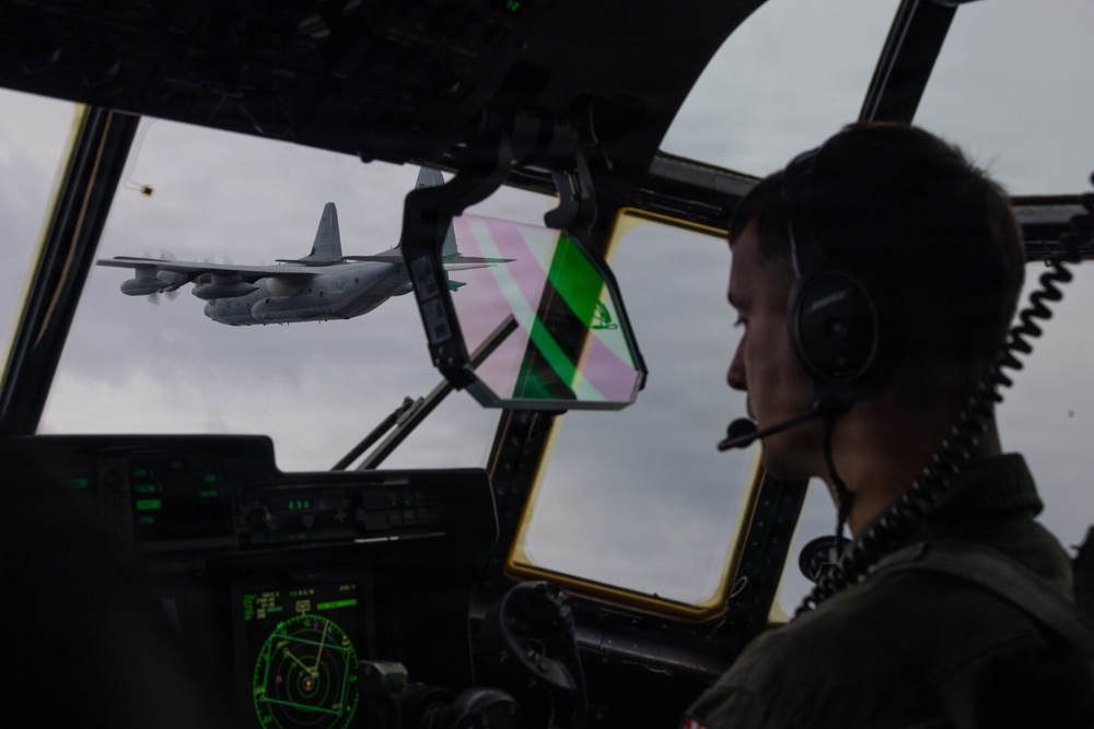 A military pilot is seated in the cockpit of a large transport aircraft, viewed from the right side, observing another similar aircraft flying in close formation outside the window. The cockpit displays multiple green-lit instrument panels and a heads-up display (HUD) with green and pink reflections visible on the windscreen. The exterior aircraft appears to be a C-130 Hercules or similar four-engine turboprop military transport.