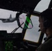 A military pilot is seated in the cockpit of a large transport aircraft, viewed from the right side, observing another similar aircraft flying in close formation outside the window. The cockpit displays multiple green-lit instrument panels and a heads-up display (HUD) with green and pink reflections visible on the windscreen. The exterior aircraft appears to be a C-130 Hercules or similar four-engine turboprop military transport.