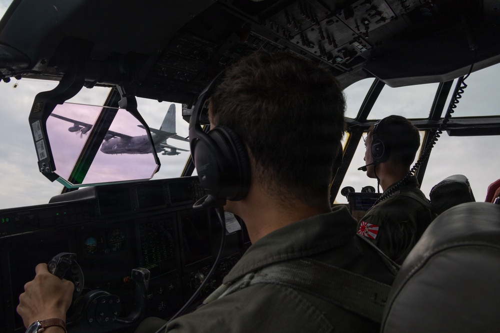 Two military aircrew members are seated in the cockpit of a military transport aircraft, flying in formation with another aircraft visible through the windscreen. One crew member displays a Japan Air Self-Defense Force (JASDF) patch featuring the Rising Sun emblem on their flight suit. The cockpit is equipped with modern avionics and instrumentation consistent with a military transport platform.