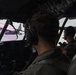 Two military aircrew members are seated in the cockpit of a military transport aircraft, flying in formation with another aircraft visible through the windscreen. One crew member displays a Japan Air Self-Defense Force (JASDF) patch featuring the Rising Sun emblem on their flight suit. The cockpit is equipped with modern avionics and instrumentation consistent with a military transport platform.