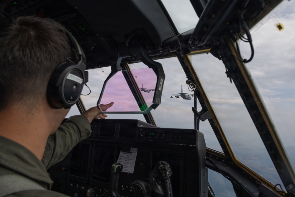 A military pilot or aircrew member is seated in the cockpit of a large military transport aircraft, pointing toward two similar aircraft flying in formation ahead. The view through the cockpit windshield reveals two turboprop military transport aircraft, consistent with C-130 Hercules variants, flying in close formation against a partly cloudy sky with a pinkish-purple horizon. The image captures an in-flight formation mission from the perspective of the trailing aircraft's flight deck.