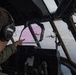 A military pilot or aircrew member is seated in the cockpit of a large military transport aircraft, pointing toward two similar aircraft flying in formation ahead. The view through the cockpit windshield reveals two turboprop military transport aircraft, consistent with C-130 Hercules variants, flying in close formation against a partly cloudy sky with a pinkish-purple horizon. The image captures an in-flight formation mission from the perspective of the trailing aircraft's flight deck.