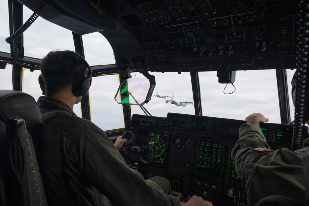 The image shows the interior of a military transport aircraft cockpit, likely a C-130 Hercules, with two flight crew members visible from behind. The pilots are wearing flight suits and aviation headsets while operating the aircraft in formation flight. Through the cockpit windscreen, at least one additional C-130 Hercules aircraft is visible flying in close formation.