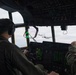 The image shows the interior of a military transport aircraft cockpit, likely a C-130 Hercules, with two flight crew members visible from behind. The pilots are wearing flight suits and aviation headsets while operating the aircraft in formation flight. Through the cockpit windscreen, at least one additional C-130 Hercules aircraft is visible flying in close formation.