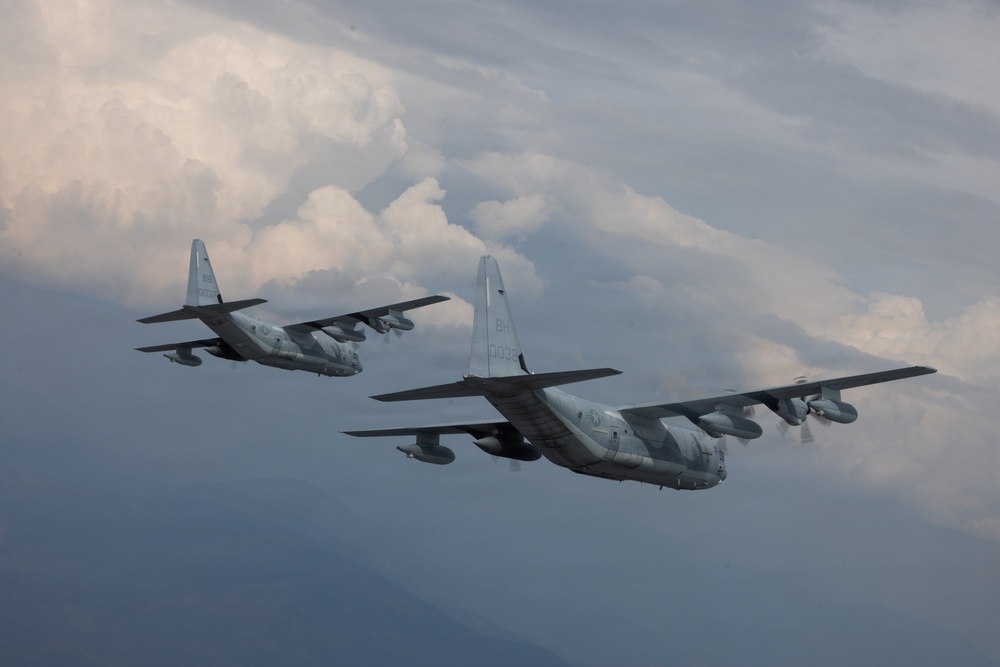 Two military turboprop transport aircraft, identifiable as C-130 Hercules variants, are flying in formation against a dramatic cloudy sky. Both aircraft are painted in gray military livery and appear to be flying at medium altitude with mountainous terrain faintly visible below. The foreground aircraft displays tail code 'BH' and serial number '0035' on its fuselage.