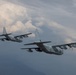 Two military turboprop transport aircraft, identifiable as C-130 Hercules variants, are flying in formation against a dramatic cloudy sky. Both aircraft are painted in gray military livery and appear to be flying at medium altitude with mountainous terrain faintly visible below. The foreground aircraft displays tail code 'BH' and serial number '0035' on its fuselage.