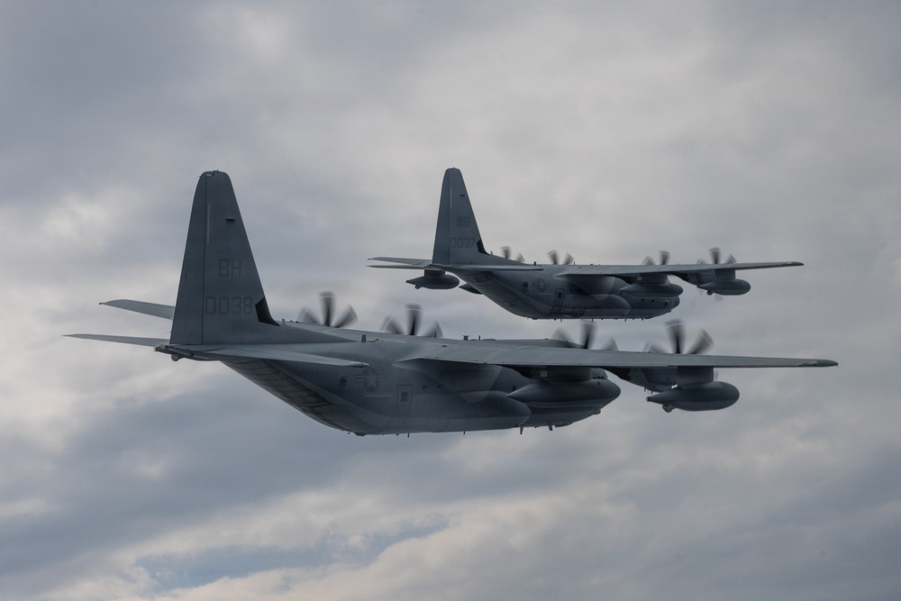 Two military turboprop transport aircraft are flying in close formation against an overcast sky. Both aircraft appear to be C-130 Hercules variants in gray military livery, with all four propellers spinning visibly. The aircraft are positioned in a staggered formation, with one slightly ahead and above the other.