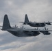 Two military turboprop transport aircraft are flying in close formation against an overcast sky. Both aircraft appear to be C-130 Hercules variants in gray military livery, with all four propellers spinning visibly. The aircraft are positioned in a staggered formation, with one slightly ahead and above the other.