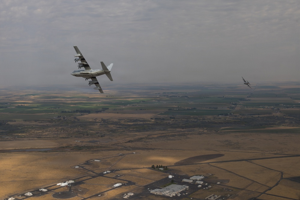 An aerial photograph showing two military aircraft in flight over a semi-arid agricultural landscape. The foreground features a large four-engine turboprop transport aircraft, identifiable as a C-130 Hercules, banking in flight. A second smaller aircraft is visible in the background to the right.