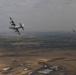 An aerial photograph showing two military aircraft in flight over a semi-arid agricultural landscape. The foreground features a large four-engine turboprop transport aircraft, identifiable as a C-130 Hercules, banking in flight. A second smaller aircraft is visible in the background to the right.