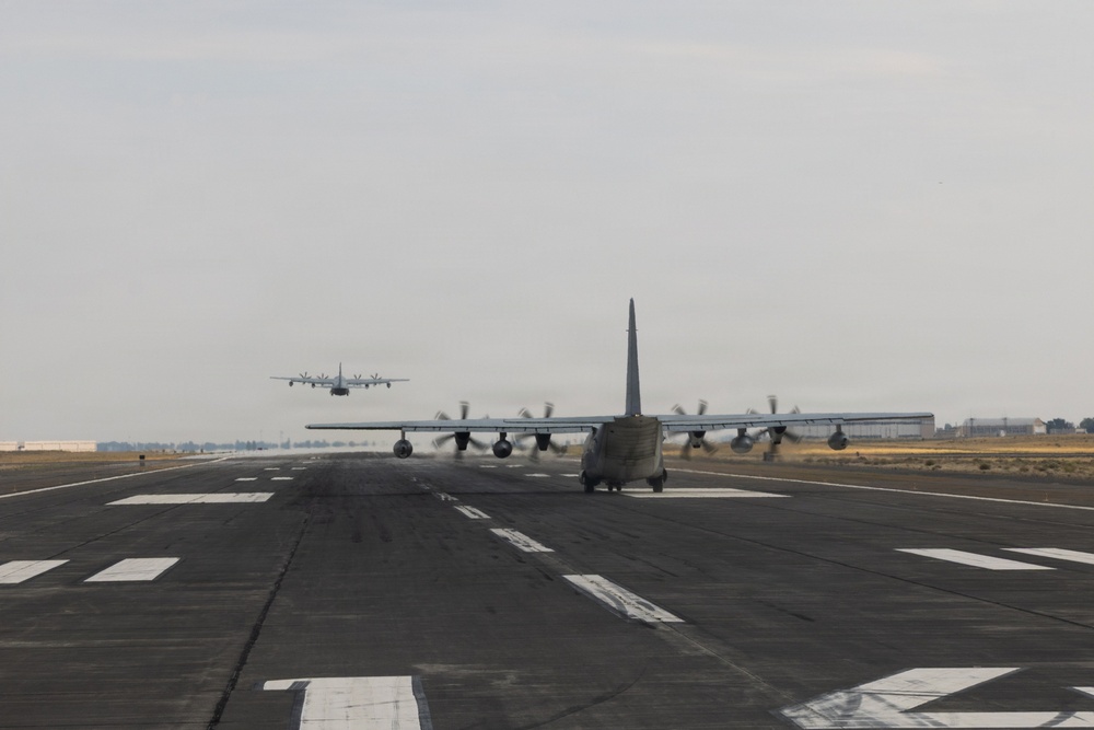 The image shows multiple military turboprop transport aircraft, consistent with C-130 Hercules variants, on and departing from a military airfield runway. At least two aircraft are visible on the ground with propellers spinning, while a third is airborne in the background. The scene appears to depict a sequential takeoff or elephant walk formation departure.