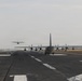 The image shows multiple military turboprop transport aircraft, consistent with C-130 Hercules variants, on and departing from a military airfield runway. At least two aircraft are visible on the ground with propellers spinning, while a third is airborne in the background. The scene appears to depict a sequential takeoff or elephant walk formation departure.