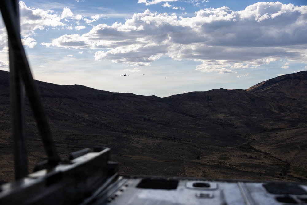 The image is taken from inside or atop a military vehicle, looking out over an arid, mountainous landscape. In the distance, two or three aircraft — likely military transport planes — are visible flying low over the ridgeline. The scene suggests a military exercise or operation in a remote, desert-like terrain.