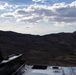The image is taken from inside or atop a military vehicle, looking out over an arid, mountainous landscape. In the distance, two or three aircraft — likely military transport planes — are visible flying low over the ridgeline. The scene suggests a military exercise or operation in a remote, desert-like terrain.
