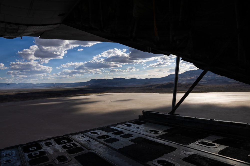 The image is taken from inside a military cargo aircraft with its rear ramp lowered, looking out over a vast desert dry lakebed or salt flat. Mountains are visible in the background under a partly cloudy sky. The aircraft appears to be on the ground or at very low altitude, with the flat terrain stretching to the horizon.