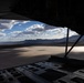 The image is taken from inside a military cargo aircraft with its rear ramp lowered, looking out over a vast desert dry lakebed or salt flat. Mountains are visible in the background under a partly cloudy sky. The aircraft appears to be on the ground or at very low altitude, with the flat terrain stretching to the horizon.