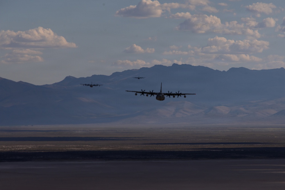 A formation of military transport aircraft, identifiable as C-130 Hercules, flying low over an arid desert landscape with mountain ranges in the background. Three aircraft are clearly visible in echelon or trail formation, with a possible fourth aircraft further in the distance. The image is taken from a low angle, likely from another aircraft or ground level, capturing the planes against a partly cloudy sky.