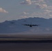 A formation of military transport aircraft, identifiable as C-130 Hercules, flying low over an arid desert landscape with mountain ranges in the background. Three aircraft are clearly visible in echelon or trail formation, with a possible fourth aircraft further in the distance. The image is taken from a low angle, likely from another aircraft or ground level, capturing the planes against a partly cloudy sky.