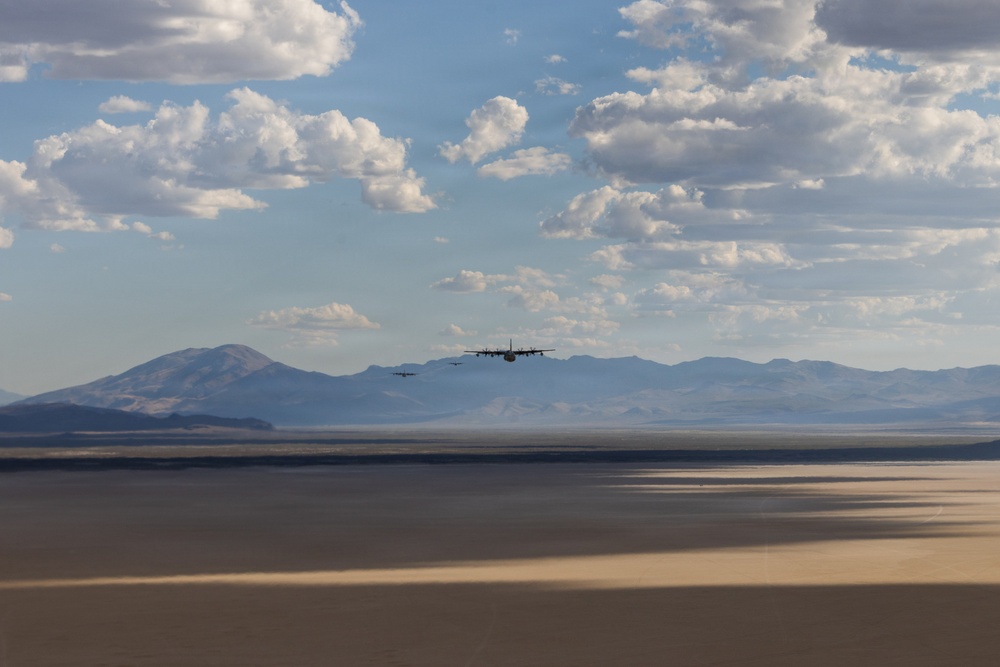Two military transport aircraft, consistent with C-130 Hercules design, fly in loose formation at low altitude over a flat desert terrain with a mountain range visible in the background. The landscape appears to be an arid basin or dry lakebed, likely in the American Southwest. The scene is photographed during daytime under partly cloudy skies with warm, diffused lighting.