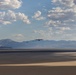 Two military transport aircraft, consistent with C-130 Hercules design, fly in loose formation at low altitude over a flat desert terrain with a mountain range visible in the background. The landscape appears to be an arid basin or dry lakebed, likely in the American Southwest. The scene is photographed during daytime under partly cloudy skies with warm, diffused lighting.