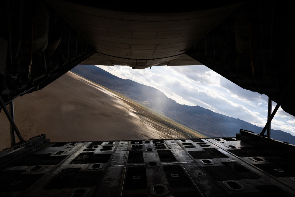 The image is taken from inside a large military cargo aircraft, looking out through the open rear cargo ramp door. A mountainous, arid landscape with a runway or taxiway is visible in the background, illuminated by sunlight breaking through clouds. The perspective suggests the aircraft is on the ground with its rear ramp lowered.