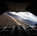 The image is taken from inside a large military cargo aircraft, looking out through the open rear cargo ramp door. A mountainous, arid landscape with a runway or taxiway is visible in the background, illuminated by sunlight breaking through clouds. The perspective suggests the aircraft is on the ground with its rear ramp lowered.